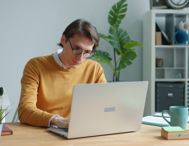 Man with glasses wearing yellow sweater looking at laptop